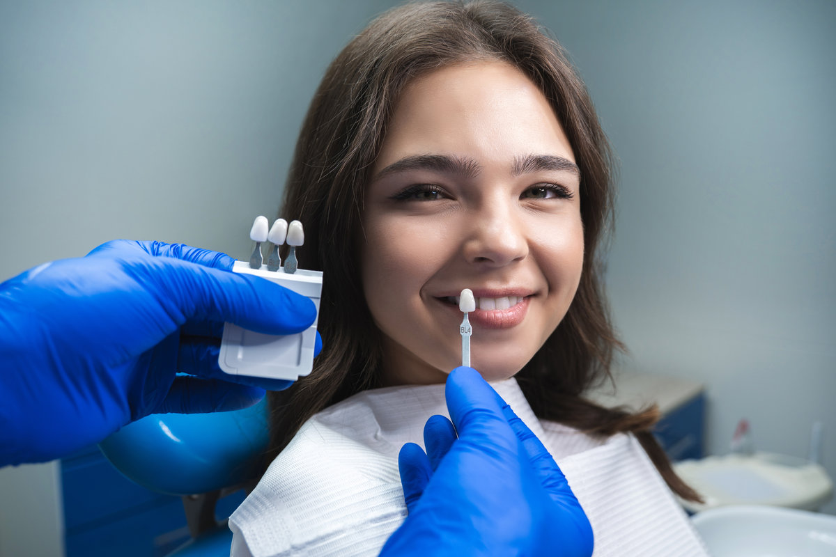 dentist in blue medical gloves applying sample from tooth enamel scale to smiling woman patient to pick up right shade for teeth bleaching procedure.