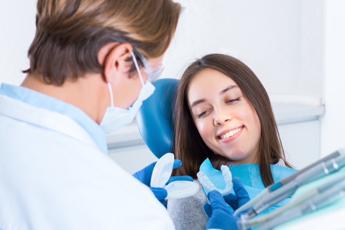 Young woman is sitting in dental blue chair in clinic, office. Man doctor is holding and teaching how to put mouth guard on teeth. Orthodontist, dentist is conducting examination of patient.