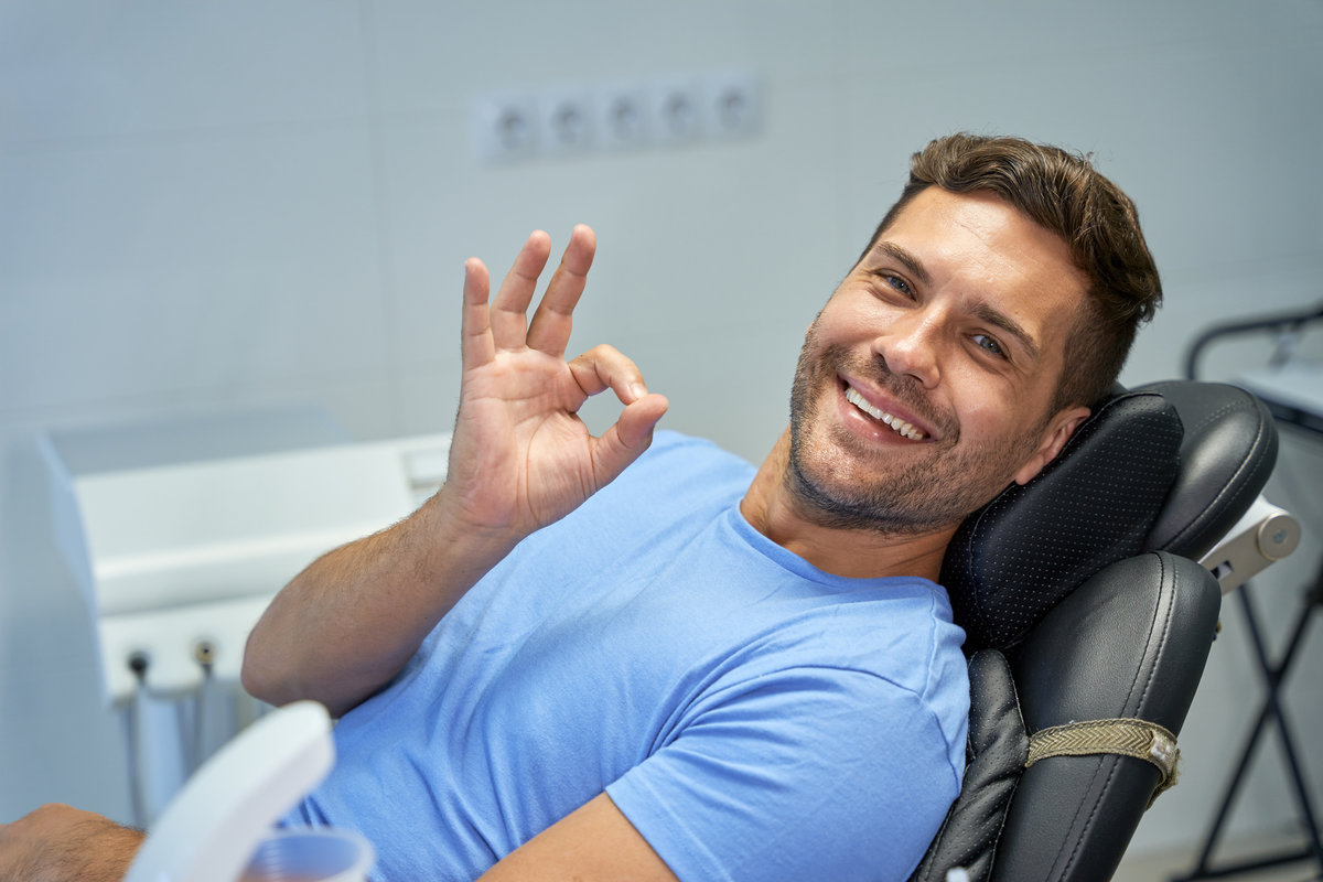 Contented young man showing an okay sign with his hand while sitting in dental chair and looking at camera