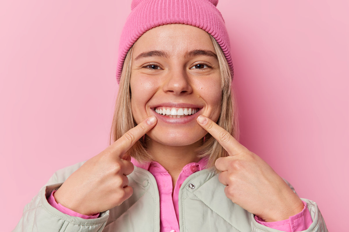 Cute positive Caucasian woman points index fingers at toothy smile shows her perfect teeth wears hat and jacket being in good mood isolated over pink background. People and happiness concept