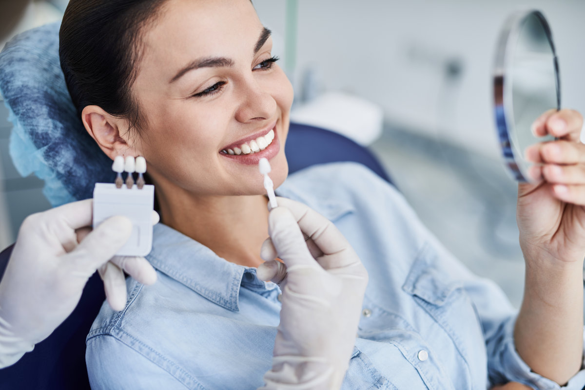 Close up portrait of charming young woman sitting in dental chair while stomatologist selecting color tone of her teeth. She is looking in the mirror and smiling