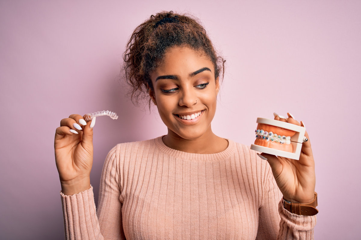 Young african american woman smiling happy holding professional orthodontic denture with metal braces and removable invisible aligner. Comparation of two dental straighten treatments